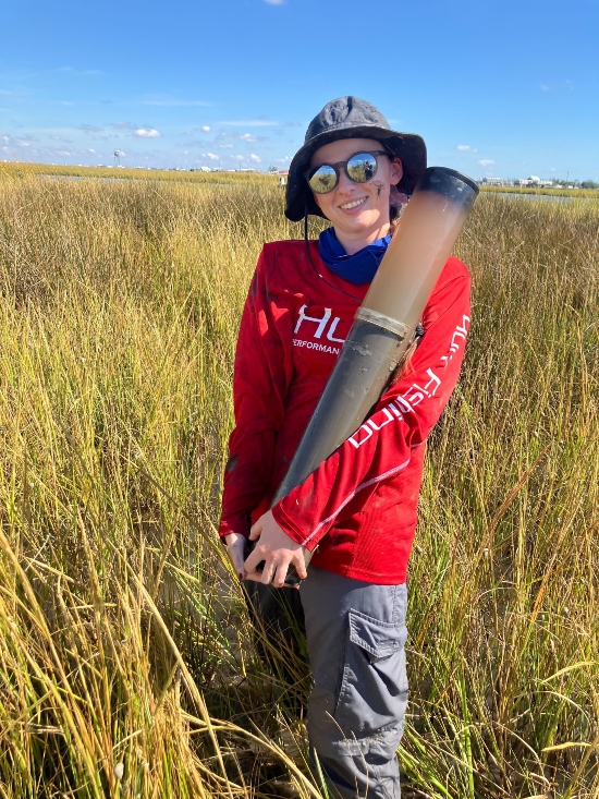 Photo shows a person wearing a red shirt, a hat, and sunglasses standing in a saltmarsh holding a tube full of mud.