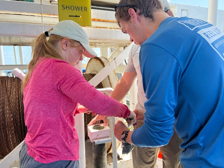 Photo show a woman in a pink shirt and a man in a blue shirt putting something in a small bag. They appear to be on a vessel.