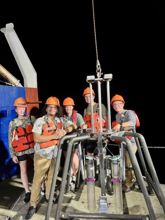 Five researchers in hard hats and life vests pose on the deck of a research vessel at night next to a sediment coring instrument used to collect seafloor samples.
