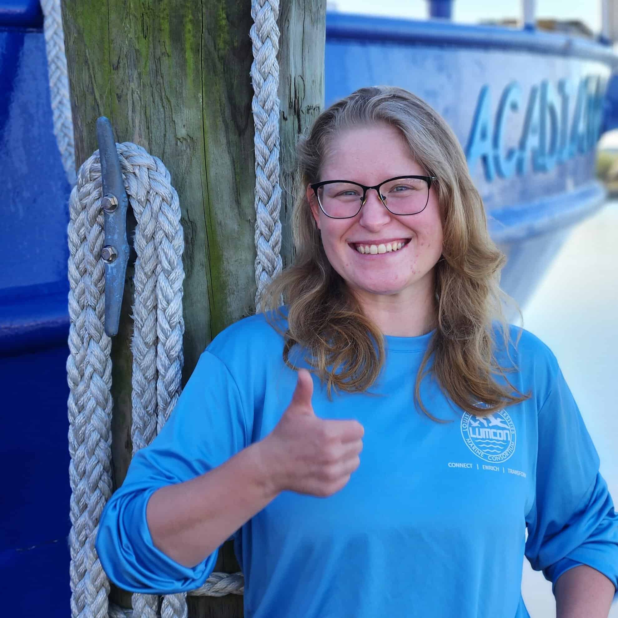 Smiling woman in blue LUMCON shirt giving thumbs up near a boat.