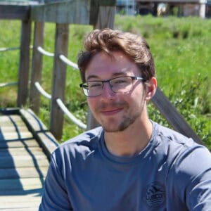 Young man with glasses smiling outdoors near a wooden railing.