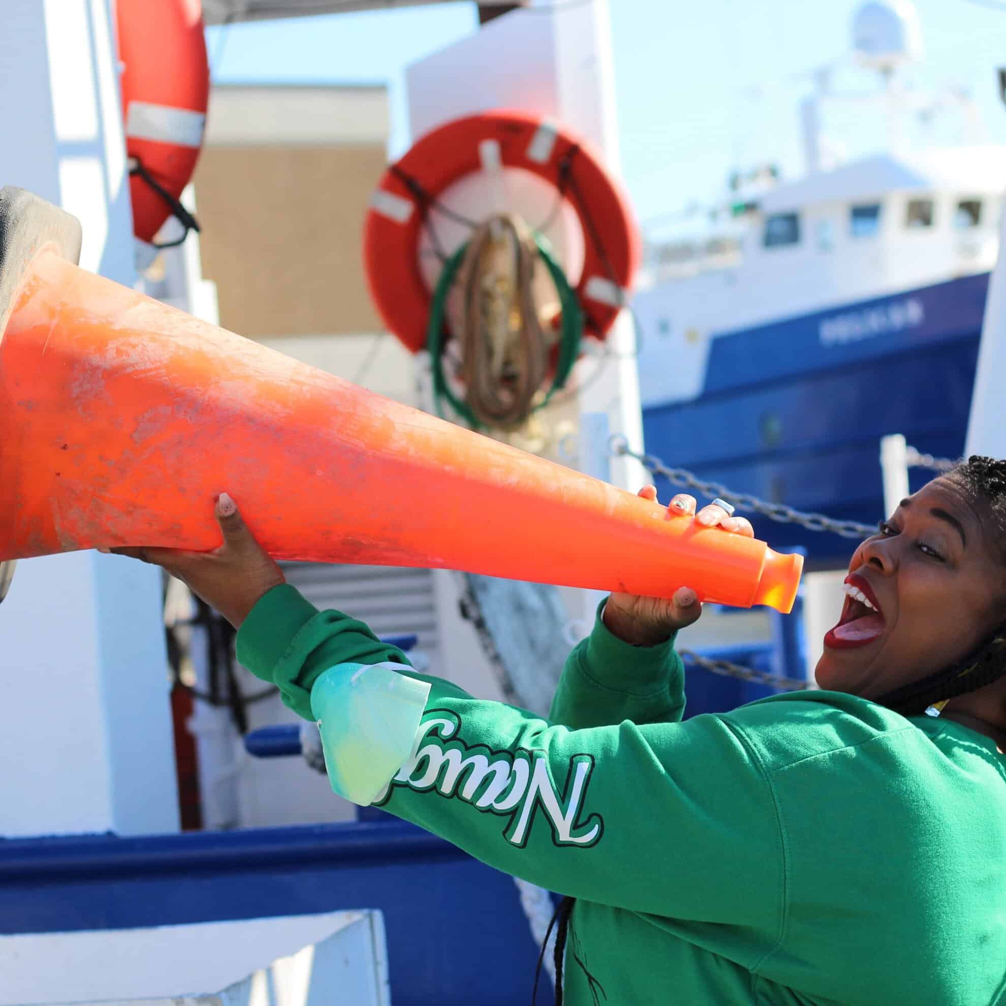 Woman in green jacket holding a traffic cone on a boat deck.