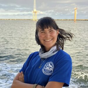 Smiling woman in blue LUMCON shirt by the water with wind turbines in background.