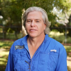 Man in blue LUMCON uniform outdoors with trees in background.