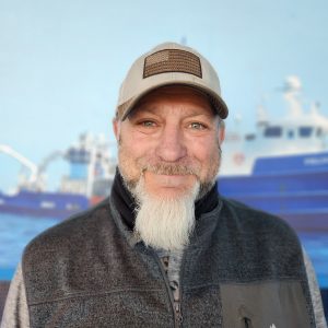 Man with a beard and cap standing outdoors near boats at sea.