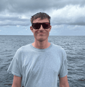 Young man in sunglasses standing by the ocean under cloudy sky.