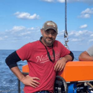 Man in red shirt on boat with ocean and blue sky background.