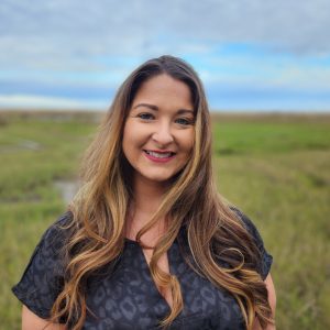 Smiling woman outdoors with a grassy landscape and cloudy sky in the background.