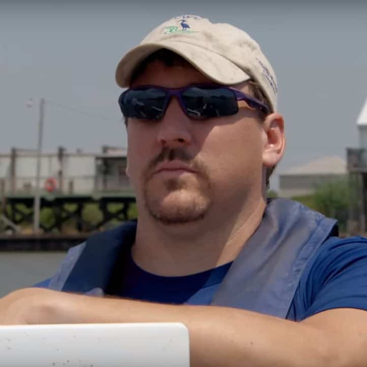Man wearing sunglasses and a cap outdoors near water.